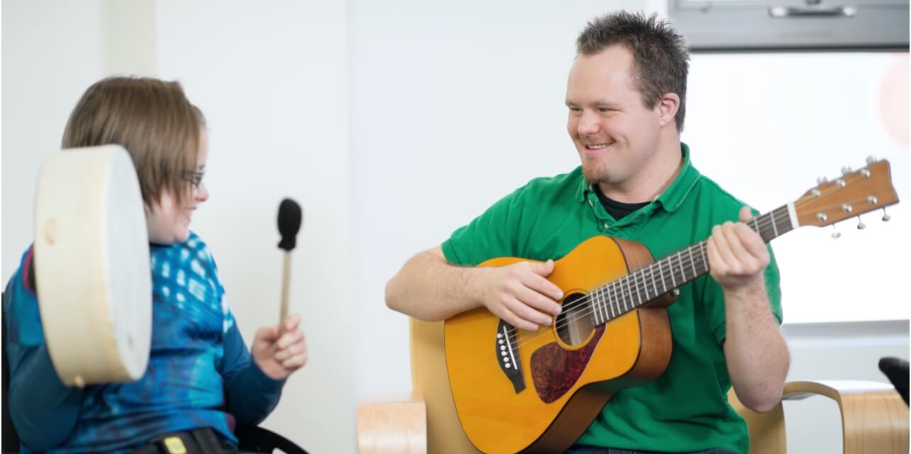 A man with a disability playing a guitar and woman playing the drums next to him