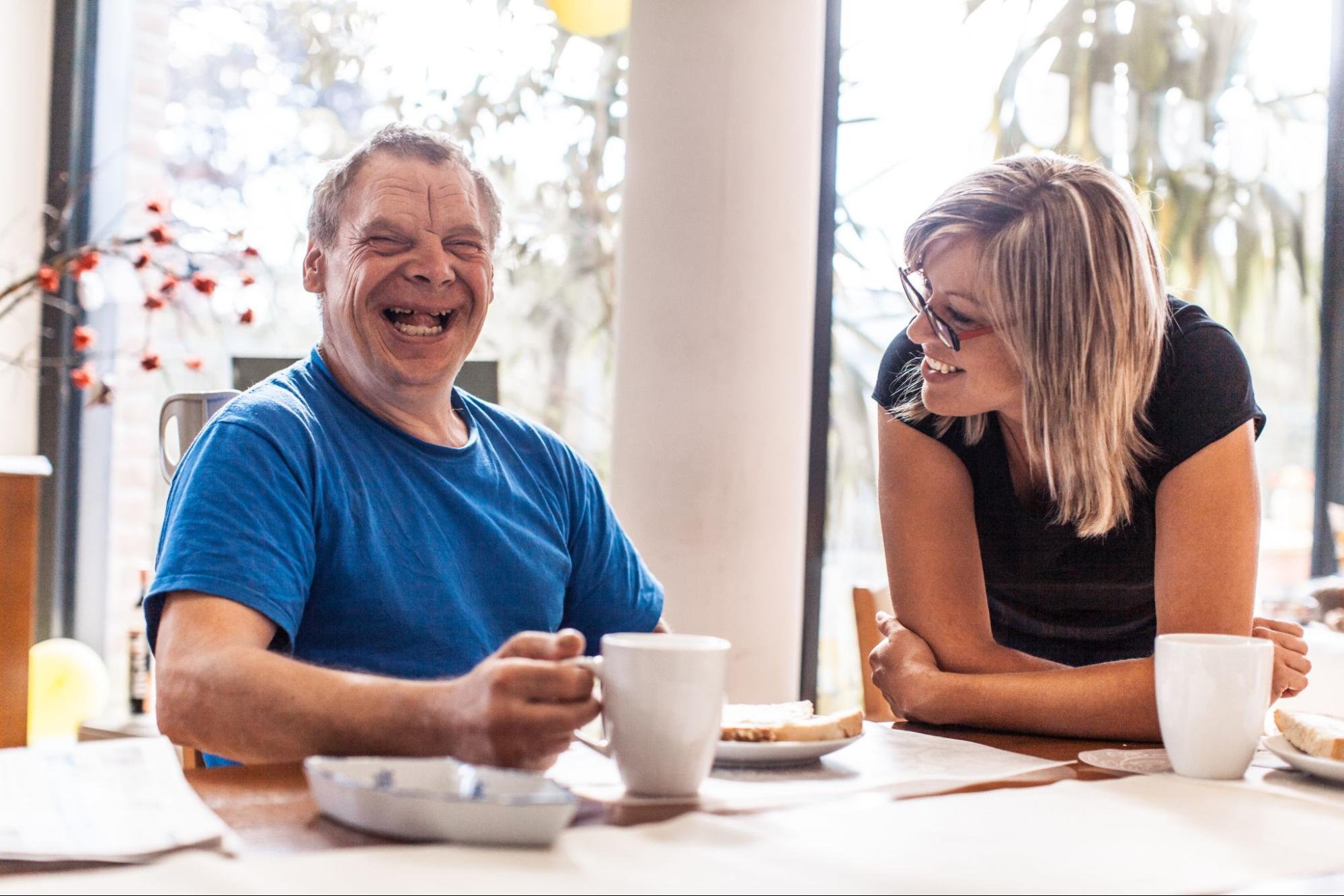 A woman sitting down smiling and looking at an older man with a disability, holding a mug