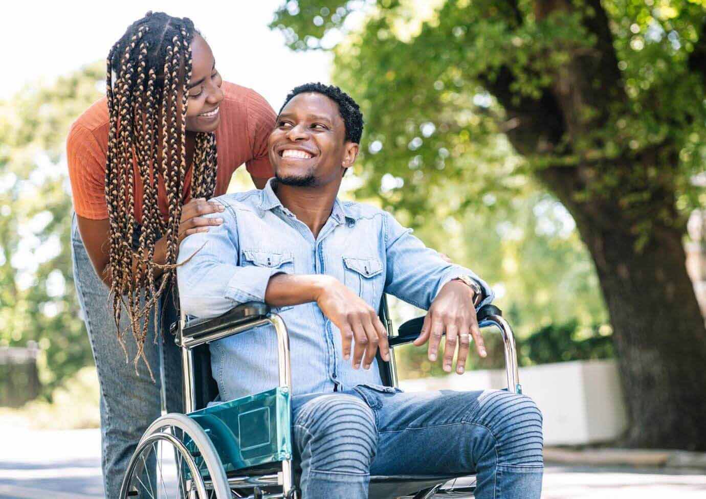 Outside, a woman stands and is leaning over behind a man who is in a wheelchair. They are smiling at each other.