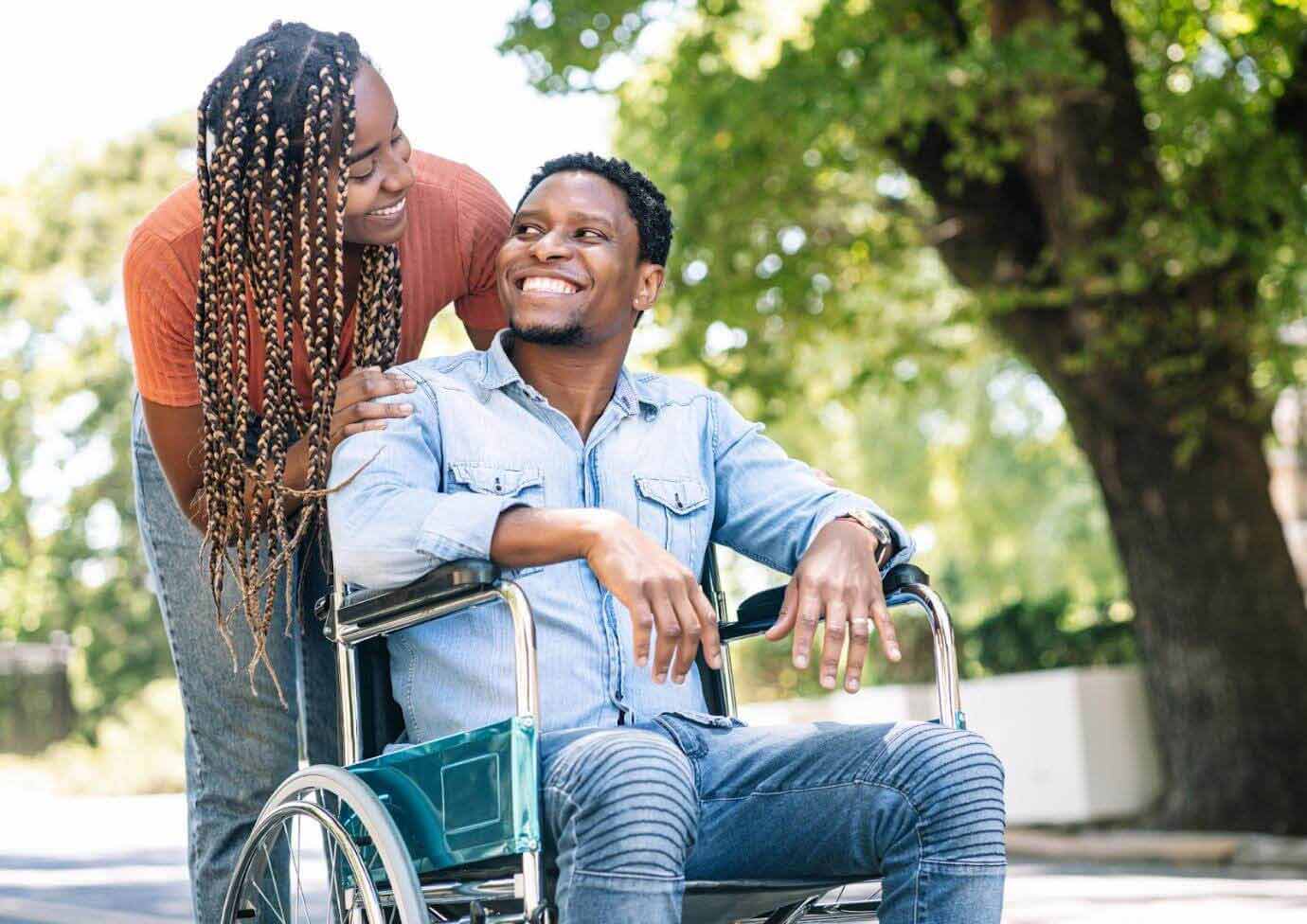 june-blog Outside, a woman stands and is leaning over behind a man who is in a wheelchair. They are smiling at each other.