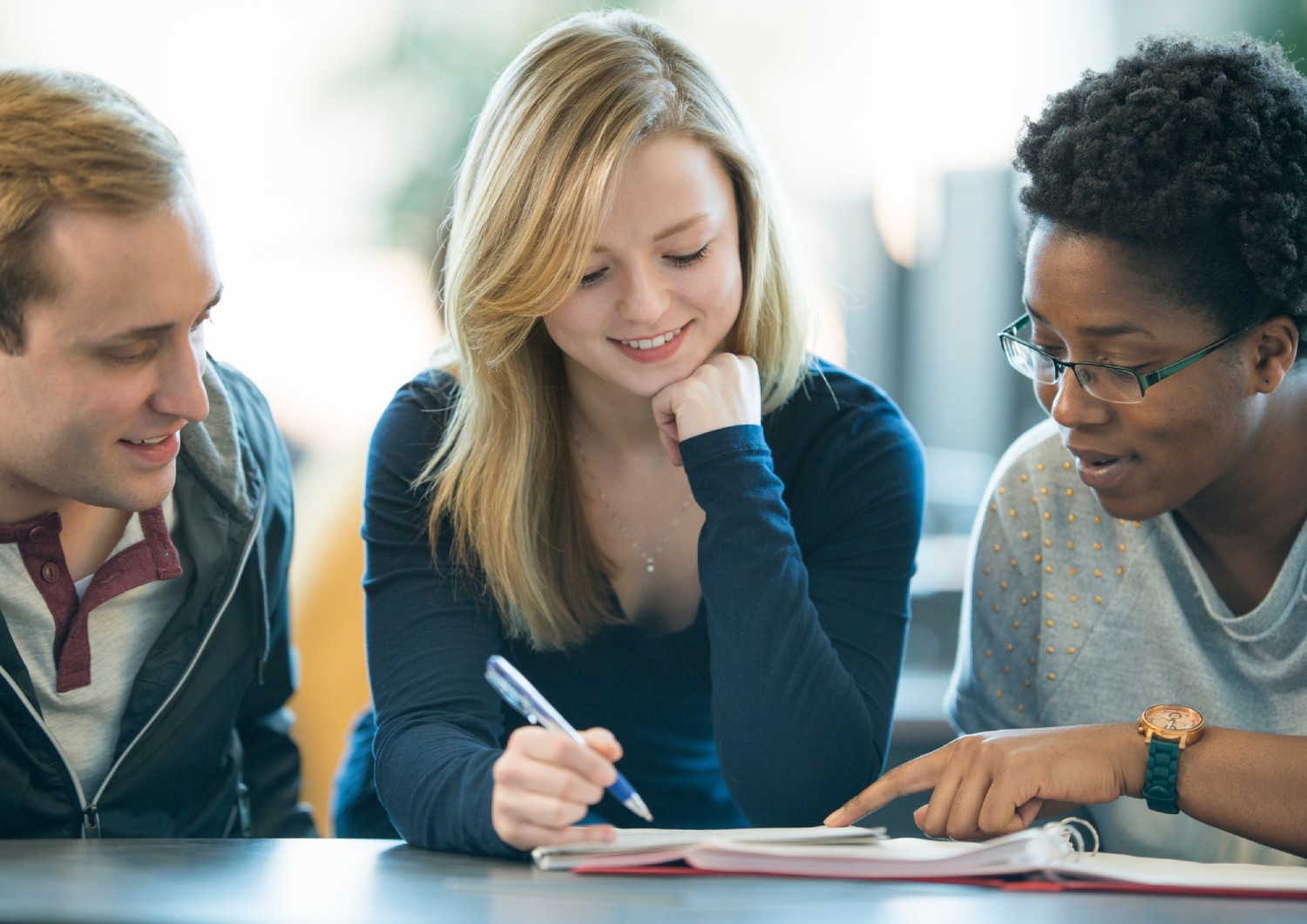 Three students sit on one side of a table studying. They are looking down at a notebook