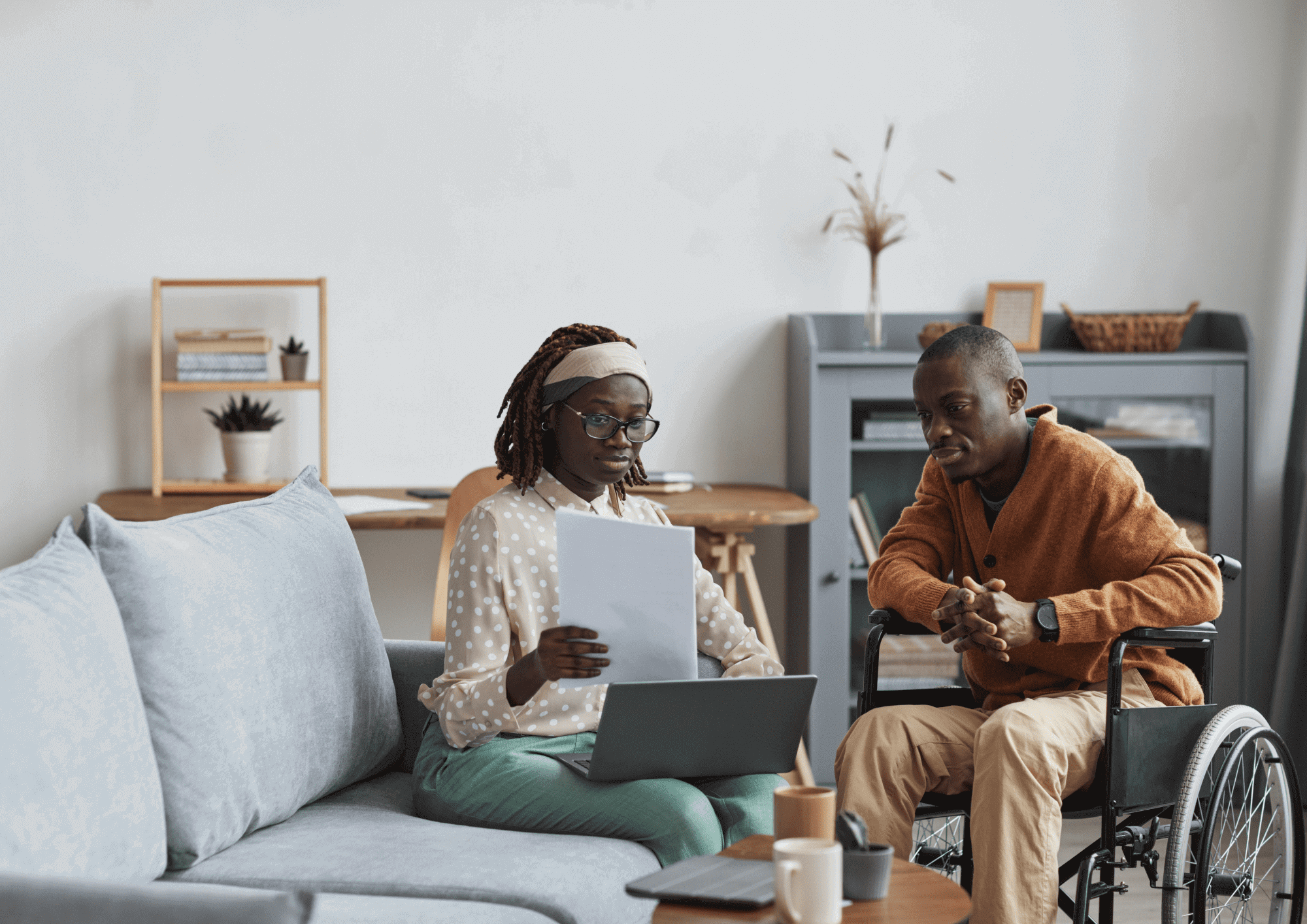 A woman sits on a couch showing a man in a wheelchair a document.