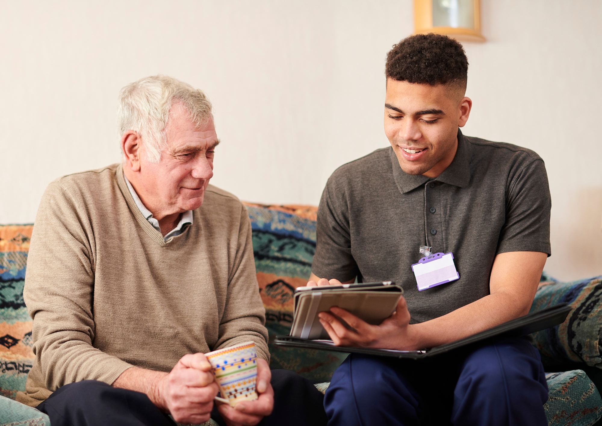 An elderly man holds a mug and sits next to a younger man who is holding an ipad.